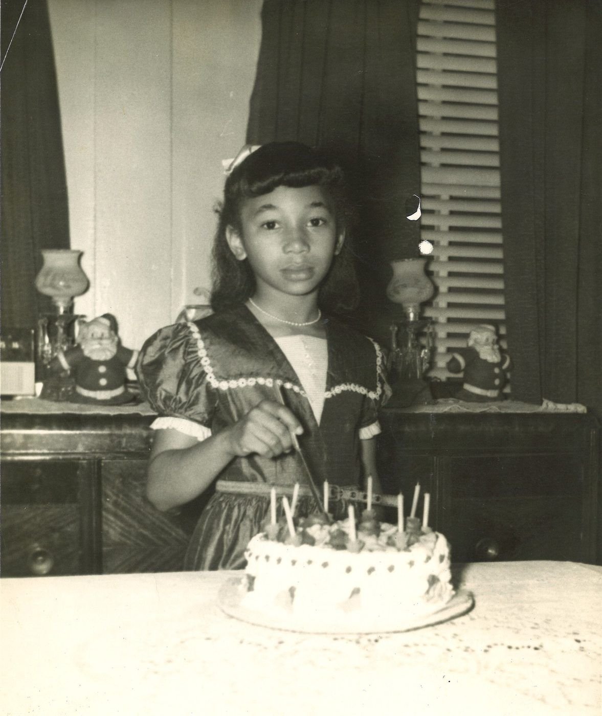 Young Carolyn in a dress and pearls cutting a birthday cake, Santa figures on the shelf behind