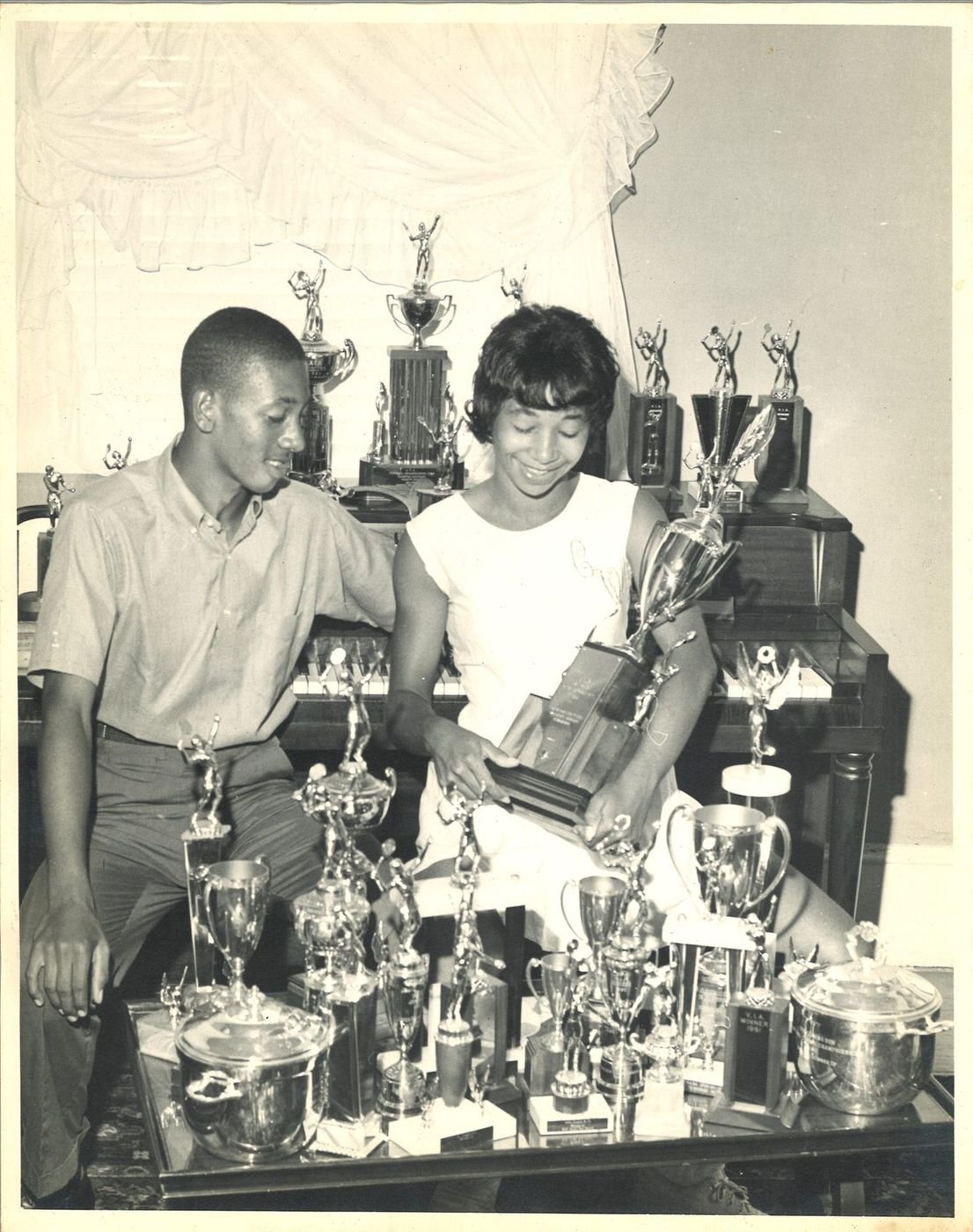 Teenage Carolyn and LaMar seated among a large collection of tennis trophies