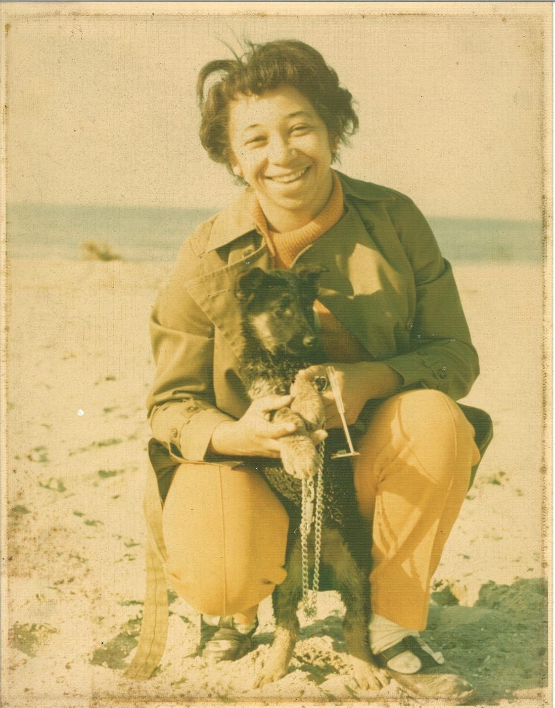 Carolyn in her twenties crouching on a beach holding a German shepherd puppy, laughing