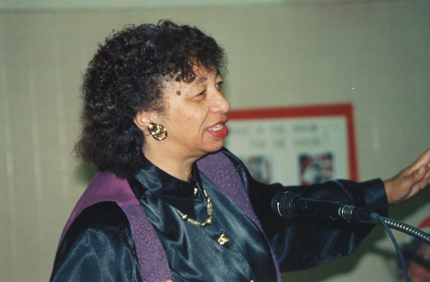 Carolyn speaking at a microphone, gesturing, in a dark satin top