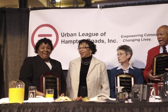 Carolyn holding an award plaque at a table with other honourees under an Urban League of Hampton Roads banner
