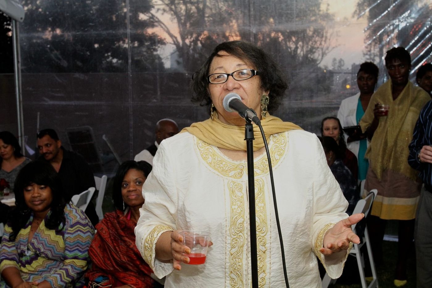 Carolyn at an outdoor evening event speaking into a microphone in a cream embroidered outfit and gold scarf