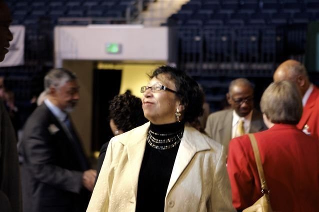 Carolyn in a cream jacket looking upward and smiling in a crowded arena