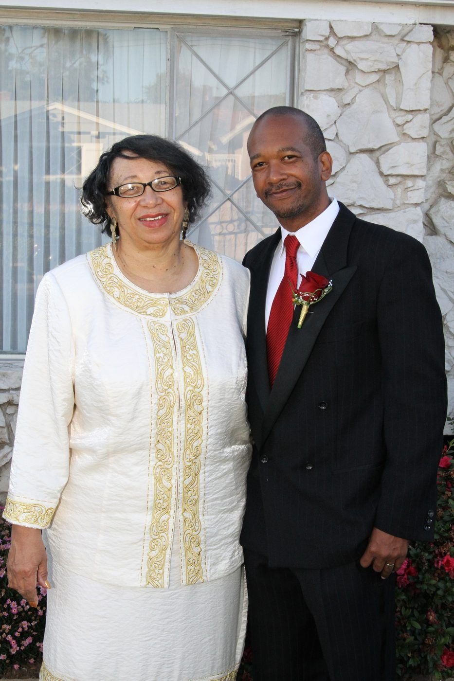 Carolyn in a cream and gold suit standing with Marcus in a black suit with a red rose boutonniere